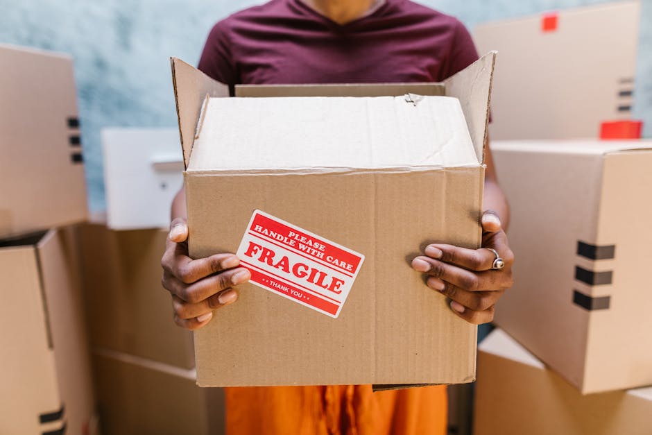 A person holding an open cardboard box labeled with a red and white 'Fragile' sticker, with their hands visible gripping the sides of the box. The individual is standing inside a room or storage area filled with additional packed boxes, some sealed with black packing tape. The background includes more cardboard cartons stacked against a wall, and the person’s torso and arms are visible, wearing a maroon shirt. The scene depicts a home relocation or furniture transport process, with the boxes prepared for moving or packing, supported by professional removals services such as those offered by Earls Court Movers, emphasizing careful handling of fragile items during the loading process.
