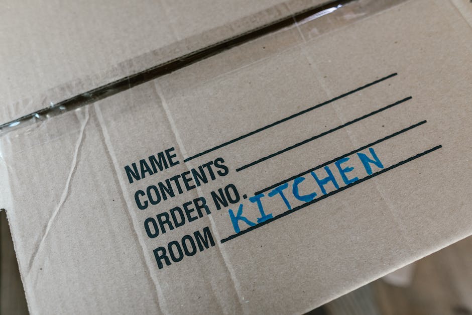 Close-up view of a cardboard moving box labeled with black and blue marker, indicating its contents and designated room for home relocation. The box is sealed with clear packing tape and appears to be placed inside a property or on a vehicle loading area, ready for transportation. The label includes spaces for name, contents, order number, and room, with 'KITCHEN' handwritten in blue on the designated room line. The setting suggests a packing and loading process as part of a house removal service, with the box prepared for transport by Earls Court Movers. The environment is well-lit, showcasing the typical materials used in moving, such as cardboard and packing tape, emphasizing the detailed organization involved in furniture transport and home relocation.