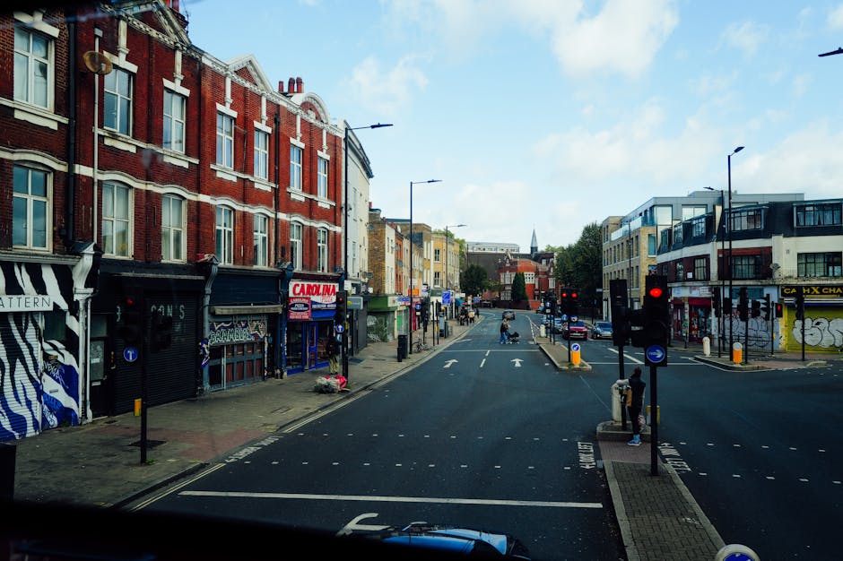 A street view of Old Brompton Road in Earls Court during daytime, showing a mix of red brick and modern buildings lining the pavement. On the left, there are closed shops with roller shutters, one with a black and white striped design, and another with a sign reading 'CARIN.' The sidewalk is active with a few pedestrians, some standing near the curb. In the foreground, the top of a vehicle's dashboard is visible, indicating the photo was taken from inside a vehicle. Across the road, on the right, there are additional commercial buildings with large windows and balconies. Traffic lights are red, and the street features designated bike lanes and road markings. The scene is well-lit by natural daylight, with a partly cloudy sky, and the overall environment reflects a typical urban setting suitable for house removals or moving logistics by Earls Court Movers.