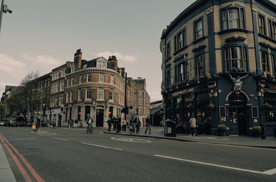 A street scene at a busy intersection in Earls Court, featuring a row of multi-story Victorian-style buildings with large windows and decorative architectural details. Pedestrians are crossing the road and walking along the pavement, some pushing bicycles or carrying bags. A black vehicle is parked near the curb, and a small group of people are gathered outside a blue pub named The Ship Wrights Arms, which has gold accents and flower boxes on the exterior. The scene is captured during daylight with soft, overcast lighting, and the overall environment reflects a typical urban setting in London suitable for house removals or furniture transport services by Earls Court Movers.