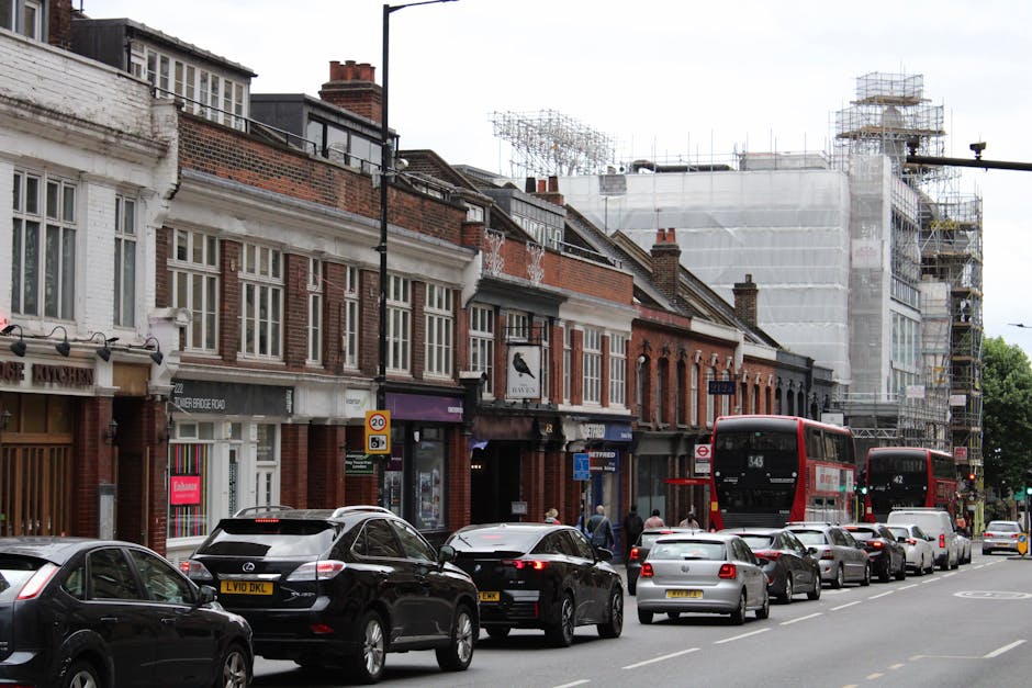 A street view of Old Brompton Road in Earls Court during daytime, showing a mix of red brick and modern buildings lining the pavement. On the left, there are closed shops with roller shutters, one with a black and white striped design, and another with a sign reading 'CARIN.' The sidewalk is active with a few pedestrians, some standing near the curb. In the foreground, the top of a vehicle's dashboard is visible, indicating the photo was taken from inside a vehicle. Across the road, on the right, there are additional commercial buildings with large windows and balconies. Traffic lights are red, and the street features designated bike lanes and road markings. The scene is well-lit by natural daylight, with a partly cloudy sky, and the overall environment reflects a typical urban setting suitable for house removals or moving logistics by Earls Court Movers.