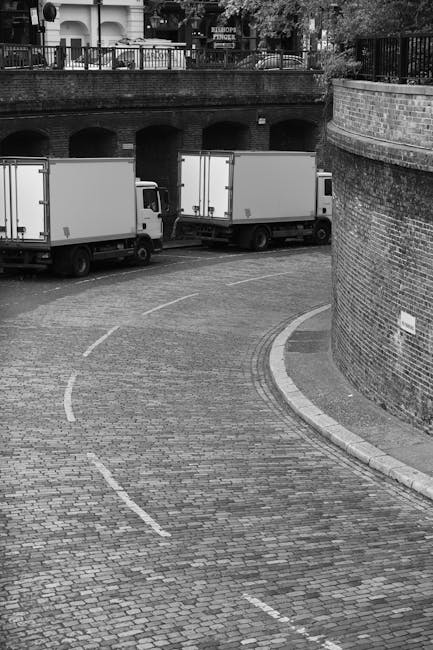 A black and white photograph depicting two medium-sized box trucks parked on a cobbled street in an urban area. The trucks, with compartmentalized cargo areas, are positioned parallel to a curved brick wall on the right side of the image, which partially encloses the street. The trucks are aligned to face toward the left, indicating they are ready for loading or unloading activities. Nearby, a set of stairs or a pathway ascends from the street, possibly leading to an upper level or property entrance. The street features faint dashed lines for traffic guidance, and the surface is constructed of brick pavers, contributing to the historic or traditional appearance of the surroundings. In the background, a brick embankment with arched openings supports a railing above, suggesting an elevated walkway or terrace area with trees and buildings visible beyond. The scene reflects a typical setting for a home relocation or furniture transport process, with suggestive elements of professional removals services as offered by companies like Earl's Court Movers, especially in the context of same-day removals and rapid availability in Earls Court.