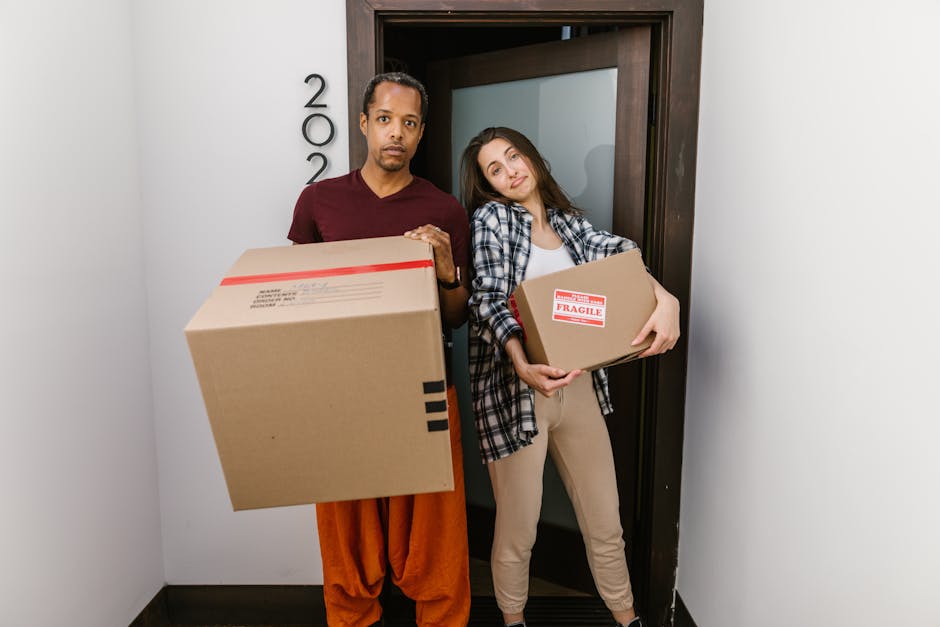 Two individuals, a man and a woman, stand inside a doorway within a residential property, each holding a cardboard box used for packing during a home relocation. The man, dressed in an orange pair of trousers and a maroon t-shirt, faces the camera with a neutral expression and holds a large cardboard box with black tape securing it, positioned in front of his torso. The woman, wearing beige pants, a white top, and a checkered shirt, leans gently against the doorframe while holding a smaller cardboard box labeled 'Fragile' with red tape, close to her chest. The doorway behind them appears to lead into an interior room, and the setting is lit with soft, natural light. This image, associated with Earls Court Movers, visually depicts the packing and loading process typical of house removal services, highlighting the careful handling of household items involved in furniture transport and moving logistics. The background includes a wooden door frame, part of the interior wall, and a glimpse of the outside pavement visible through the open door, emphasizing the transition from inside to outside during the moving process.